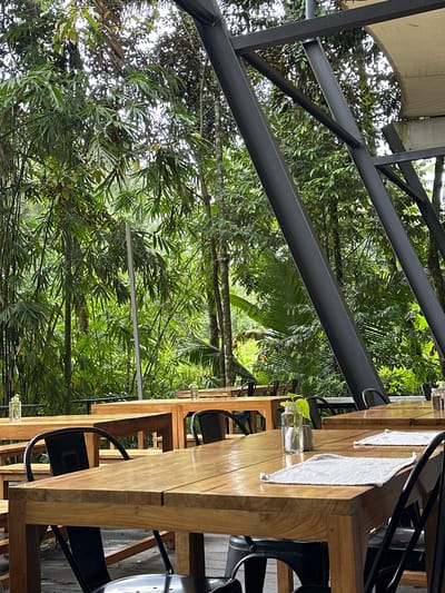 The image shows a restaurant in Licin, Indonesia. The restaurant has wooden tables and black chairs set up for dining. On the tables, there are napkins and small vases with plant cuttings. The restaurant is open-air, with a metal roof structure and a backdrop of lush green vegetation, including trees and bamboo.