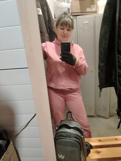 A woman is captured in a mirror selfie indoors, wearing a pink hooded tracksuit. She has light-colored hair with bangs and a slight smile, holding a black smartphone in her hand which is covered by a black glove. The background, visible in the mirror's reflection, reveals a functional space featuring grey metal lockers. Cardboard boxes are stacked on top of the lockers, with faint text including "FESTIV" visible on one box. A black jacket hangs on the right side of the frame. To the left of the mirror, a white paneled wall is seen. At the bottom of the image, a dark grey backpack with a prominent metallic letter 'W' is placed on a light wooden bench. The setting appears to be a changing room or storage area, located in Fedorino, Russia.