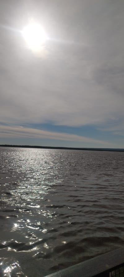 The image shows a body of water under a sky with a bright sun obscured by clouds. The water surface reflects sunlight, creating shimmering patterns. The horizon line is visible with a dark strip of land in the distance. The location cannot be determined from the image. No people, interactions or other objects are visible.