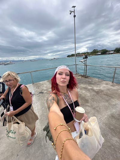 Two women are on a concrete pier in Kampung Toyapakeh, Indonesia. One woman has red hair, a white headband, and tattoos on her arm. She wears a black top and gold bracelets. She is holding a coffee cup and a white bag. The other woman is blonde and wears glasses, a black top, a khaki skirt, and sandals. She also holds a white bag. The sea is in the background with some boats and the sky is cloudy.
