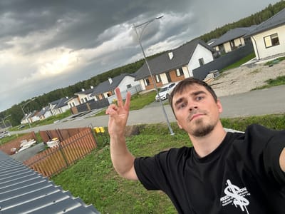 A man is taking a selfie outdoors in a new residential development in Rassokha, Russia. He is wearing a black t-shirt and making a peace sign with his left hand. Behind him, a row of modern houses with dark gray roofs are visible. The sky is overcast with dramatic dark clouds. A street lamp is positioned between the man and the houses. A wooden fence and green grass are in the foreground.