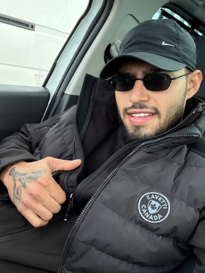 A male individual is depicted from the chest up, seated inside a vehicle in Gateshead, United Kingdom. He is wearing a black Nike baseball cap with a white swoosh logo, and dark oval-shaped sunglasses. He has a beard and is smiling slightly, revealing his teeth. His attire includes a black puffer jacket over an inner black garment, possibly a fleece or additional jacket. The puffer jacket features a circular "ZAVETTI CANADA" patch on the right chest area. His right hand, visible in the foreground, has a grey/black tattoo of a stylized animal (possibly a bird or creature) on the back. This hand is positioned near the central zipper of his jacket. The interior of the vehicle shows a white door pillar on the left, with a window revealing a blurry, light-colored exterior and unreadable text on the glass. The subject is looking directly at the camera.