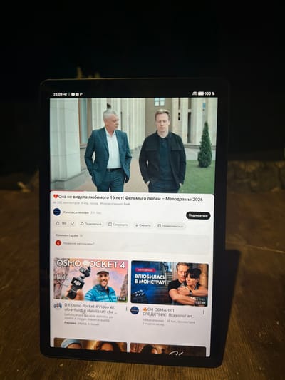This is a media file captured from a tablet, displaying a YouTube interface. The tablet is placed on a wooden surface, suggesting an indoor setting, possibly a home or cafe. The lighting is dim, with the tablet screen being the primary light source, creating a contrast with the dark surroundings.

The screen displays a YouTube feed with several video thumbnails. The main video in focus is a thumbnail for a film titled "Она не видела любимого 16 лет! Фильмы о любви – Мелодрамы 2026". It features two individuals, a man and a woman, in a dramatic pose, with a tag that reads "МЕЛОДРАМА". This video has accumulated 46,235 views and was posted 4 weeks ago by "Киновселенная". There are also options to like, dislike, share, save, and download the video, along with a comment section.

To the right, another thumbnail is visible for a video titled "ОН ОБМАНУЛ СЛЕДСТВИЕ! Психолог вл...", which appears to be a psychological thriller. Further down, a third thumbnail advertises "DJI Osmo Pocket 4 Video 4K ultra-fluidi e stabilizzati che...", a product review or demonstration related to camera technology.

At the bottom of the tablet screen, a status bar shows the time as "23:09", indicating it's late in the evening. The battery level is at 100%, and Wi-Fi is connected. The overall mood conveyed by the video thumbnails leans towards drama, mystery, and technology. The tablet's orientation is portrait, and it's held at an angle, suggesting it's being used by someone who is either watching or browsing content.