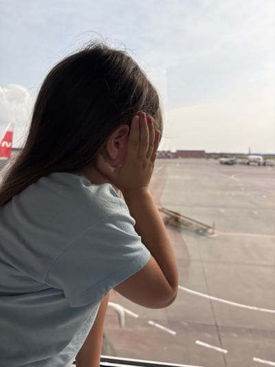 A young girl, viewed from behind, leans against a window, her left hand covering her ear. She wears a light blue t-shirt. Her dark hair cascades down her back. The window looks out onto an airport tarmac. Several airplanes are visible in the distance, along with airport vehicles and buildings. The sky above is bright and hazy. The scene is likely at an airport in or near Bol'shie Kabany, Russia, given the context.