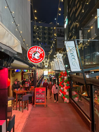 This image depicts a brightly illuminated commercial pedestrian alleyway at night in Saint Julian's, Malta. String lights with warm white bulbs crisscross overhead, complemented by large illuminated white star decorations.
On the left side of the alley, a red circular sign for "NATIVE BAR MALTA'S HOTTEST LAYUP BAR" is prominently displayed. Below it is an outdoor bar seating area under a white awning, furnished with multiple wooden high-top tables and dark chairs. Approximately seven individuals are seated at these tables; in the foreground, one man in a dark jacket is observed looking at a mobile phone. Overhead heaters are visible within this area. A red sandwich board sign on the ground advertises "HAPPY HOUR BUY 1 GET 1 FREE" and "NIBBLES HAPPY HOUR 50% OFF."
On the right side, multiple white vertical banners featuring the "Bacc by Hugo's" logo are suspended. Below these banners, a glass-enclosed area, likely an outdoor seating extension of a restaurant, shows tables and chairs inside. A large arch or garland constructed from red, white, and green balloons stands on this side, extending towards the foreground.
The central pedestrian path is paved. Four individuals are walking in the same direction, from the background towards the foreground. These include a man in a red top and yellow beanie, a woman in a beige jacket, a man in a grey zippered sweatshirt, and another man in a dark jacket.
In the background, modern multi-story buildings with balconies and illuminated windows rise on both sides of the alleyway.
