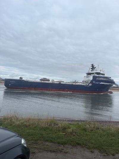 A large offshore support vessel, the "REM FORTRESS," registered in LONGVA, is depicted transiting the River South Esk in Montrose, United Kingdom. The vessel, colored dark blue with a red hull, is moving from left to right, creating a visible wake. Its white and blue superstructure features "REM Offshore" branding and the identifier "PSV 06 CD." The sky is entirely overcast with a uniform layer of grey, rippled clouds. In the immediate foreground, a portion of a dark-colored vehicle, including its headlight and part of the bonnet, is visible on the lower-left. The shoreline consists of patchy green and dry brown grass, dirt, and scattered small rocks and pebbles. The far bank of the river shows indistinct low-rise structures, consistent with port or industrial facilities. No individuals are present.