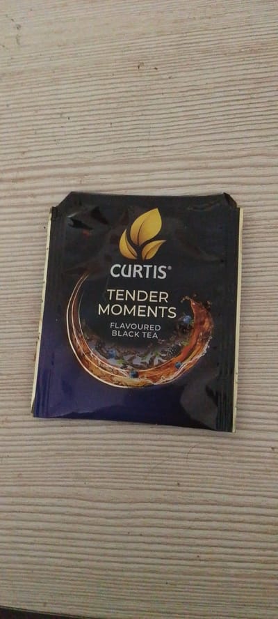 The image shows a single tea sachet, branded "CURTIS TENDER MOMENTS FLAVOURED BLACK TEA". The sachet is dark blue with a gold leaf logo and white text. It is positioned centrally on a light-colored wooden surface with a distinct horizontal grain pattern. The lighting suggests an indoor environment. There is no indication of the city of Lyudinovo, Russia, or any context related to it in the image. The focus is solely on the tea sachet and the wooden surface it rests upon.