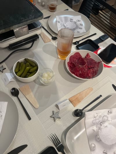 This media file captures a meticulously set dining table for a raclette meal, likely an evening gathering in Bellevue, Switzerland. The table features light grey placemats and is equipped for multiple diners.
Prominently displayed is a raclette grill with a black cooking surface, central to the meal. Accompanying food items include a large white oval bowl filled with thin slices of reddish cured meat. Another white bowl contains green gherkins, and a small glass holds pickled pearl onions, all common raclette accompaniments.
Each place setting is equipped with an oval light grey plate, a white paper napkin adorned with a subtle grey pattern of baubles, and black cutlery including a fork, knife, and spoon. Small black raclette pans are positioned next to some settings, along with individual wooden spatulas. Clear ribbed glasses are present, one holding an amber-colored drink.
Decorative touches include small silver glittery star ornaments and white place cards or labels, with "Claire" and "Mélanie" visible on two of them. Mobile phones are also seen on the table. Although no people are visible, the arrangement suggests a prepared social event or dinner. The indoor lighting indicates that the meal is taking place in the evening.