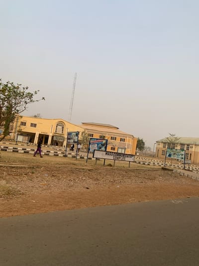 A hazy daylight scene displays a beige, multi-story building marked "MASS COMMUNICATION COMPLEX" with prominent signage. A tall radio tower rises behind it. To the right, a tan grassy area meets an asphalt road. Another similar beige building stands further back. A pedestrian walks along a striped curb. The sky is a muted, overcast color.