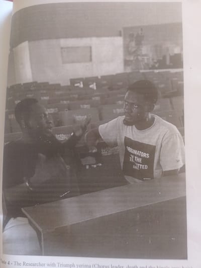 This black and white photograph captures an indoor scene, likely a lecture hall or auditorium, with rows of seating visible in the background. Two young men are the central subjects, seated at a table in the foreground. The man on the left, wearing a dark shirt, is gesturing with his hands as if speaking or explaining something. The man on the right, wearing a t-shirt with text, is looking towards the man on the left with a focused expression.

The t-shirt of the man on the right has visible text that reads "ORIGINATORS OF THE", with the word "LITTED" appearing below, possibly part of a larger word or phrase. The word "SINCE" is also visible. Another visible text element appears on a dark object held by the man on the left, reading "tettung". Below the image, a caption is partially visible, reading "Plate 4 - The Researcher with Triumph yerima (Chorus leader, death and the king's gray hair)". This suggests the individuals are potentially involved in a research project or performance, and the context might be related to a cultural or artistic event. The lighting suggests an indoor setting, possibly during the daytime, though no specific time of day or weather conditions can be determined. The overall atmosphere appears to be one of focused discussion or instruction.