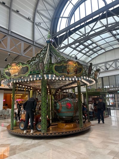 The image captures an indoor carousel located in a shopping center or mall environment. The carousel is ornately decorated with painted scenes, lights, and mechanical elements. People are present, some standing near the carousel and others potentially riding it. The background reveals architectural elements such as a glass ceiling with a network of steel beams. Based on the presence of "Porte de Serris" signage, the location can be identified as Serris, France. There is a "SWEET FOOD NAISE" restaurant in the background. The floor is tiled and appears clean.