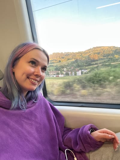 A young woman with dyed pink and purple hair is sitting by the window of a train. She is wearing a purple fleece hoodie and gray sweatpants. Her left hand is resting on her lap, and her right hand is visible near the bottom of the frame. The train is moving through a rural area with rolling hills, green fields, and scattered houses. The sun is setting, casting a warm glow on the landscape. The location in the city of Llanera, Spain is not discernible from the image.