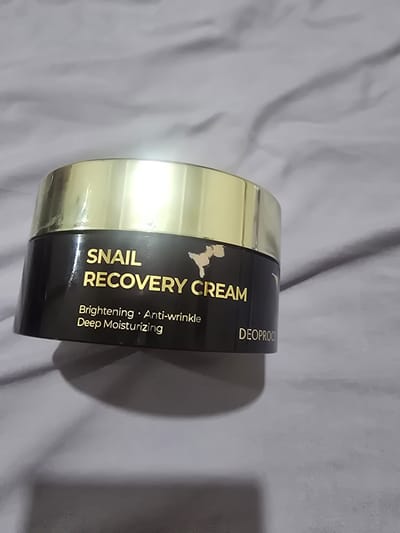 A dark jar with a gold lid sits on a grey, wrinkled surface. Gold lettering on the jar reads "SNAIL RECOVERY CREAM," and below that, "Brightening • Anti-wrinkle • Deep Moisturizing." Further down the jar, "DEOPROC" is visible in gold lettering. The lid of the jar is reflective and shows a bright light source. The background is a textured grey fabric that appears to be a blanket or bedsheet, with visible folds and shadows. The object appears to be a cosmetic product. The location is not discernible from the image.