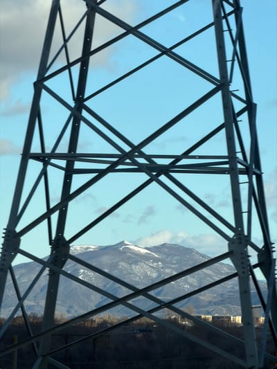 The image displays an outdoor scene in Akhalsopeli, Georgia. The foreground is dominated by the dark gray, bolted lattice structure of a metal power transmission tower, which extends across the entire frame, from bottom to top and side to side. Visible through the angular openings of the tower's framework is a mid-ground landscape featuring a range of mountains, partially covered with white snow, under a clear blue sky interspersed with scattered white clouds. The most prominent mountain peak in the center is heavily snow-covered. Below the mountain range and above a dark, indistinct band of foreground vegetation, a series of light-colored, low-rise buildings are discernible, indicative of a settlement. No individuals or specific actions are depicted.