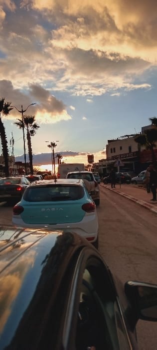 A street scene with cars lined up along the right. A blue and white Dacia is in the middle. Palm trees and buildings line the left side of the street, silhouetted against a sunset sky filled with clouds. The foreground features part of another car, obscuring the view. The overall perspective is tilted, creating a slightly disorienting effect.
