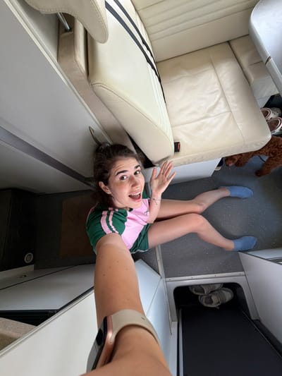 A young woman with dark hair is sitting on the floor of what appears to be a camper van or recreational vehicle, taking a selfie. She is wearing a pink and green t-shirt and blue socks. Her legs are extended in front of her, and she is smiling and waving at the camera. A small brown poodle is visible in the background, partially obscured by the woman's legs and the seat. The interior of the vehicle is clean and organized, with light-colored seats and white cabinets. The floor is a speckled grey material. There is a shoe storage compartment visible below the seats containing a pair of sneakers. The scene appears to be indoors, possibly in the city of Nea Moudania, Greece, based on the context provided.