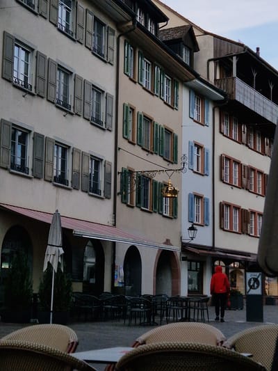 The image depicts a building in Rheinfelden, Switzerland, characterized by rows of windows with varying shutter styles and colors, including green, blue, and brown. A prominent street lamp with ornate metalwork hangs from the building's facade. The lower level of the building features arched entryways, one of which leads to a shop or restaurant. An open umbrella stands in front of one of the archways. On the sidewalk, there are tables and chairs typical of an outdoor café or restaurant. A person wearing a red jacket is visible walking on the sidewalk. A no-parking sign is also visible near the pedestrian.