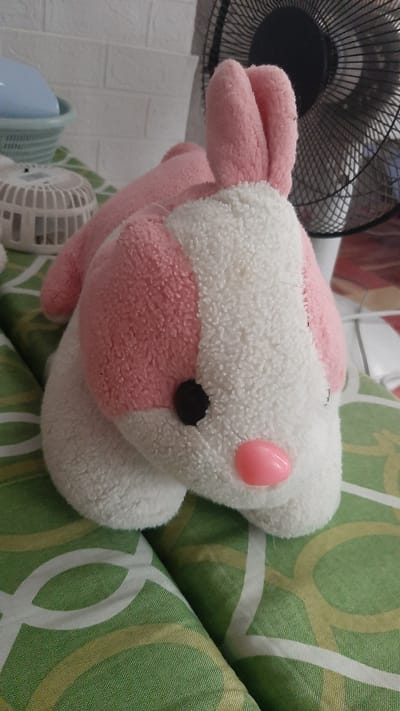 A pink and white plush bunny toy is positioned on a green and white patterned surface, likely bedding. The bunny has large pink ears, a white fluffy body, a pink nose, and a black eye. In the background, a black oscillating fan is visible, along with a white textured wall and a blue object within a light blue container. To the left of the bunny, a small white fan-like object with a cage-like structure is also present. The scene appears to be indoors, possibly in a bedroom. The specific location in Kawit, Philippines, cannot be determined from the visual information provided.