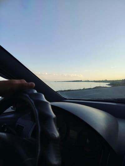 The media file shows a first-person perspective from inside a car, driving along a road adjacent to a large body of water. A hand is visible on the steering wheel, indicating the driver. The scene is set during the daytime, likely late afternoon or early evening, given the soft, warm light and clear sky with some scattered clouds.

The car's dashboard is visible in the foreground, with the steering wheel and part of the instrument panel in shadow. The view through the windshield encompasses a calm body of water, possibly a lake or sea, stretching towards the horizon. Along the far shore, a distant treeline and some buildings, including what appears to be construction cranes, suggest human habitation or development. The road the car is traveling on curves alongside the water, with grassy verges and a single white line marking the lanes.

The overall impression is one of a peaceful, scenic drive. The expansive sky and the tranquil water create a sense of openness and calm. The location context provided is Bulan-Segettu, Kyrgyzstan, which aligns with the visual cues of a large body of water and potentially developing infrastructure. There is no visible text in the media file.