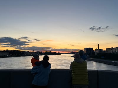 This image depicts a family enjoying a sunset over a river in Staraya, Russia. In the foreground, silhouetted figures of people are visible from behind, facing the water and the cityscape. A woman is holding a baby on her left shoulder, and another person stands to her right, also facing the sunset. The baby is wearing an orange jacket and a gray hat. The woman is wearing a blue denim jacket. The person to her right is wearing a yellow puffer vest over a gray hooded sweatshirt. The river occupies the middle ground, reflecting the warm colors of the sky. The background shows the silhouette of buildings and trees lining the riverbank, with a tall smokestack visible on the right side of the frame. The sky is a gradient of soft blues and oranges, with some clouds scattered across it, indicating the end of the day. The overall impression is one of a serene family moment against a beautiful natural backdrop in a Russian city.