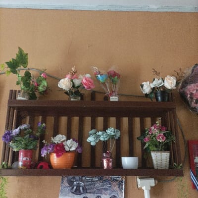 The image depicts an indoor scene, likely within the city of Pasir Muncang, Indonesia, showcasing a wall-mounted two-tiered dark brown wooden shelf unit adorned with various artificial flower arrangements. The wall is painted a light tan or peach color, with a white ceiling border visible at the top.
On the upper shelf, from left to right, are: a clear glass container holding green artificial foliage with small pink flowers; a small pot with white and pink artificial roses; two individual bouquets of artificial flowers (one blue and red, one predominantly red) wrapped in clear plastic with ribbons; and a dark pot containing white and cream artificial flowers. To the far right, a large bundle of pink artificial flowers is partially visible.
The lower shelf, from left to right, displays: a red can, possibly repurposed, holding purple and green artificial flowers; an orange ribbed ceramic pot with white and dark pink artificial flowers; a clear pink-tinted jar containing light blue/green artificial flowers on thin stems; a plain white ceramic cup; and a light-colored woven basket pot with pink and green artificial flowers and foliage. Green artificial foliage trails off the right edge of this shelf.
Below the lower shelf, a printed poster or paper with text including "18" 18X27 EXCEL" and "ACE," along with an image of a dark object and a batik-like pattern, is affixed to the wall. To its right, a white rectangular device, possibly a security camera or sensor, is mounted on the wall with visible wiring. No individuals are present in the scene.