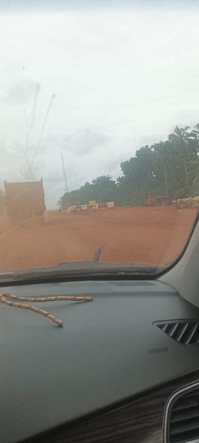 The image shows a view from inside a car, looking out onto a dusty, reddish-brown road. A large truck is visible on the left side of the road. Stacks of what appear to be bricks or similar materials are lined up on the right side. In the background, there are green trees and what appears to be a construction crane. Two thin, wrapped sticks are visible on the dashboard of the car. The word "AIRBAG" is visible on the car's dashboard. The location is Oto, Nigeria.