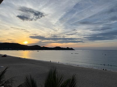 The image depicts a serene sunset scene at a beach in Song Cau, Vietnam. The foreground is dominated by a sandy beach, with the silhouettes of palm fronds visible at the bottom edge. The middle ground features the gentle waves of the ocean lapping onto the shore, with a few small boats anchored in the distance. Several people are present on the beach and in the water: two individuals are walking along the shoreline, two are sitting on the sand, and a group of about five people are visible in the shallow water, seemingly swimming or wading. The background showcases a chain of dark, silhouetted hills or mountains against a sky filled with dramatic clouds. The sun is setting on the left side of the frame, casting a warm, golden glow on the horizon and reflecting on the calm sea. The overall atmosphere is peaceful and tranquil.