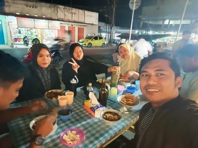The image depicts a group of people gathered around a table at what appears to be an outdoor eatery in Luwoo, Indonesia, during nighttime. The foreground is dominated by a man taking a selfie, smiling at the camera. He is wearing a black jacket with white stripes. To his left, another man is partially visible, also looking towards the camera.
The table is covered with a blue and white checkered tablecloth and is laden with food and drinks. There are bowls of food, cups, a bottle of dark liquid, and a tissue box. Several other individuals are seated around the table, some of whom are wearing headscarves. One woman in a black hijab is making a peace sign with her fingers, while another woman in a yellow hijab is holding a woven container. In the background, there are buildings with illuminated signs, motorcycles, and a yellow car. The overall atmosphere suggests a casual dining experience.