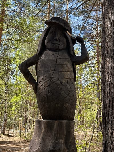 The image shows a wooden sculpture of a human figure with outstretched arms, located in what appears to be a wooded area. The sculpture is dark in color and has a rough, textured surface with some evidence of carving and cracking. The figure is surrounded by trees and foliage. Based on the prompt, the location is Sayansk, Russia.