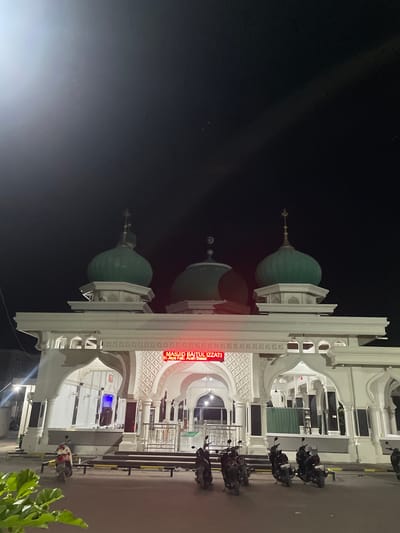 In the city of Siron, Indonesia, a mosque named Masjid Baitul Izzati stands illuminated at night. The mosque features three prominent green domes, with the central dome being the largest. The facade is white and ornate, with arched entrances and decorative patterns. A red neon sign above the main entrance displays "MASJID BAITUL IZZATI" and below it, "In Jaya Kab. Aceh Besar". Several motorcycles are parked in front of the mosque on the paved ground, which is bordered by yellow and black striped bollards. A person in red is visible near one of the motorcycles. The sky above is dark, indicating nighttime. Lush green foliage is present in the lower left corner of the image.