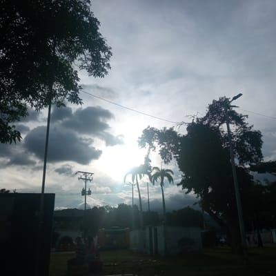 This image captures a cloudy sky over El Limón, Venezuela, with the sun partially obscured by dark, dramatic clouds. The scene is dominated by silhouetted trees and palm trees in the foreground and midground. A tall, thin pole with a street lamp is visible on the right, and another pole with power lines and a transformer is seen to the left. In the distance, a hilly landscape is visible beneath the clouds. The lower portion of the image shows a park-like area with a dark rectangular structure on the far left, followed by some low buildings, a green inflatable structure, and two individuals seated. The overall lighting suggests either early morning or late afternoon.