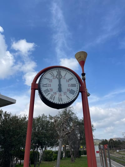 The image displays an outdoor public clock situated at a railway station in Buñol, Spain. The central object is a large, circular clock with a white face, featuring black Roman numerals (I-XII) and an inner ring of red Arabic numerals (13-23). Both the hour and minute hands are positioned vertically, indicating 12:00. This clock is supported by a red, arched metal frame, which rests on two vertical red poles. Attached to the upper section of the right pole is a two-toned, conical public address speaker, red at its base and fading to a lighter color at its wider opening, with a small dark camera or sensor mounted directly beneath it.
To the left of the clock, a partial view of a light-colored overhead structure, possibly an awning or building extension, is visible. The ground area beneath and between the poles is covered with green grass, interspersed with various green bushes and several trees, some with dark foliage and others appearing deciduous with gnarled, bare branches. To the right, a paved platform with discernible markings extends into the midground, beyond which multiple railway tracks are visible running horizontally. The background features rolling hills with sparse vegetation, set against a bright blue sky scattered with white cumulus clouds. No individuals or active events are present in the scene.