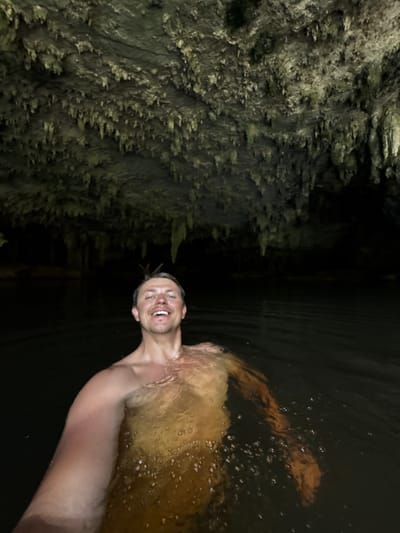 A man with short brown hair, wearing no shirt, is submerged in dark water within a cave. He is smiling and looking directly at the camera. The cave ceiling is visible above him, covered in stalactites. The water appears murky and the surrounding environment is dark, suggesting an underground location. The image was taken in Ao Luek, Thailand.