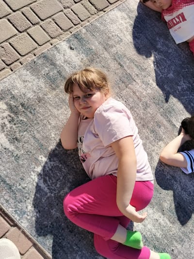 This overhead shot captures two young girls lying on a rug outdoors, with a paved brick area visible to the left. The girl in the foreground, with light brown hair, is lying on her side, propped up on her arm and hand. She wears a light pink t-shirt and bright pink leggings, with green socks on her feet. She looks directly at the camera with a slight smile. To the right, another girl, partially visible, lies on her back. She is wearing a white t-shirt with "BROOKLYN" and "NEW YORK" printed in red text, and pink shorts or a skirt. Shadows cast by unseen objects suggest it is daytime, with bright sunlight. The setting appears to be a residential area or public space in Staryi Aidar, Ukraine.