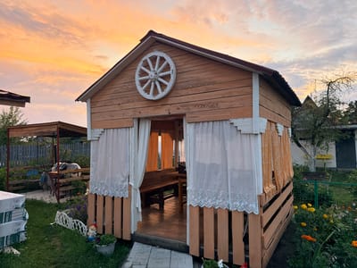 In the city of Радужскі сельскі Савет, Belarus, at the golden hour of sunset, a wooden gazebo stands as the central focus. The gazebo, constructed from light-colored wooden planks, features a gabled roof with a dark red corrugated metal covering. A decorative white wagon wheel is affixed to the front gable. White curtains with intricate lace trim hang from the opening, framing a view of wooden benches and a table inside. The gazebo's base is made of horizontal wooden slats, and it is situated on a paved area.
Surrounding the gazebo, a vibrant garden blooms with flowers in shades of purple, yellow, and orange. To the left of the gazebo, a stack of white, wrapped construction materials is visible on a patch of green grass. A small white fence segment and a garden gnome are placed in the grass near the gazebo's entrance. Further back, another covered structure with a sloped roof can be seen, partially obscured by foliage. The background includes a gray wooden fence, trees, and a white building with a dark door and a yellow bench. The sky is filled with dramatic, colorful clouds painted in hues of orange, pink, and yellow, indicative of a sunset.
