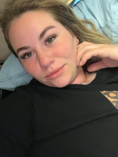 A close-up, high-angle selfie of a young woman with blonde hair, light green eyes, and fair skin with freckles. She is lying down on a light blue pillow and a blue blanket, with her head tilted. Her left hand is touching her cheek and chin. She is wearing a black t-shirt with a leopard print detail on the right side. She has applied false eyelashes and appears to be wearing subtle makeup. The overall lighting is soft and natural. The image does not contain any visual information that allows for the determination of its location within Vyatskiye Polyany, Russia, or any other city.