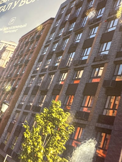 The image shows a modern apartment building in Kozulino, Russia. The building features a dark grey brick facade with regularly spaced windows, many of which are dark or reflect the sky. Some windows have subtle red or orange accents visible within them. A brown brick building is visible to the left, appearing older or a different architectural style. The lower portion of the image is partially obscured by a vibrant green tree with bright yellow leaves, suggesting it is either spring or summer. A streetlamp with a cream-colored shade is also visible in the lower left. The text on the upper left corner indicates "ПОДЗЕМНЫЙ ПАРКИНГ" (underground parking) and a parking symbol, suggesting the building has this amenity. The overall impression is of a contemporary urban residential area.