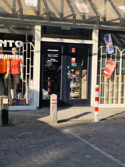 This media file captures the exterior of a retail store specializing in sports and teamwear, possibly named "NTO" and "FRANKEN." The store has a glass storefront, with mannequins displaying athletic apparel, including orange and red shirts. Promotional items like Coca-Cola and Red Bull cans are displayed on the exterior and inside the window. The store's opening hours are posted on the door: Tuesday to Friday from 09:30 to 18:00, and Saturday from 09:30 to 16:00. Signs for DPD pickup and "Flesjes parkeren" are also visible.

The scene appears to be set during the daytime, with sunlight casting shadows on the pavement, suggesting it might be late afternoon. The weather seems clear and sunny. The location is identified as Schaijk, Netherlands, with the store address possibly indicated by "8E" on the facade. The overall atmosphere is that of a commercial establishment open for business, with no people directly visible in the frame. The focus is on the storefront, its displays, and promotional materials.
