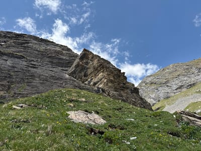 The image depicts a steep, grassy mountainside with scattered rocks and a large boulder prominently featured in the foreground. The terrain slopes upwards towards imposing rock faces. The sky above is a clear blue with scattered white clouds. A thin wire stretches across the upper portion of the image, disappearing out of frame, and a small, square metal structure is visible on the ridge of the left-hand rock face. The environment suggests a natural, mountainous landscape. This scene is located in the vicinity of Leukerbad, Switzerland, likely on a hiking trail or in an alpine region. There are no people or animals visible in the image.