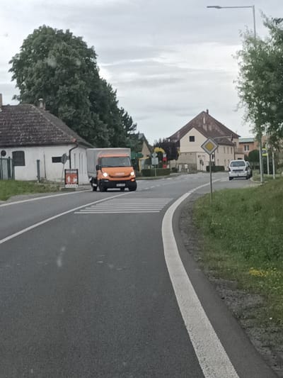 The image depicts a scene in Dřevčice, Czechia, on an overcast day. A grey, cloudy sky suggests it might be morning or afternoon, with no strong sunlight. The setting is a rural road with a white lane marking curving to the right.
In the foreground, an orange Iveco truck is driving over a pedestrian crossing. The truck has its headlights on, and the word "IVECO" is visible on the front. Behind the truck, on the left side of the road, is a white building with a dark tiled roof, appearing to be a house or small business. A red and white sign with figures is mounted on a pole in front of this building.
Further down the road, on the right, a white van is parked. Beyond the van, there's a prominent yellow diamond-shaped traffic sign on a pole. Several houses with tiled roofs are visible in the background, nestled amongst trees. Green grass and trees line the sides of the road, with some yellow wildflowers visible on the right. A street lamp with a modern, elongated light fixture stands tall on the right side of the frame. The overall atmosphere is calm and ordinary, suggesting a typical day in a small Czech village.