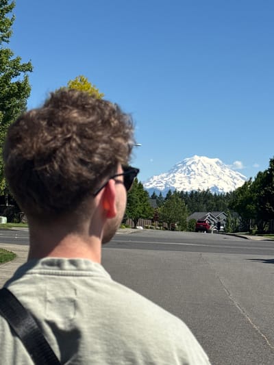 A medium shot captures the back of a young man wearing sunglasses and a light gray t-shirt, facing a snow-capped mountain range in South Hill, United States of America. The man's curly brown hair is visible, as is the strap of a dark bag draped over his shoulder. In the background, a paved road curves through a residential neighborhood lined with trees and houses. A red SUV is parked in front of one of the houses. The sky is clear and blue, and the sun is shining brightly, casting shadows on the road. The mountain range is majestic and imposing, dominating the horizon with its snow-covered peaks. The overall impression is one of a bright, sunny day in a peaceful suburban setting with a striking natural backdrop.