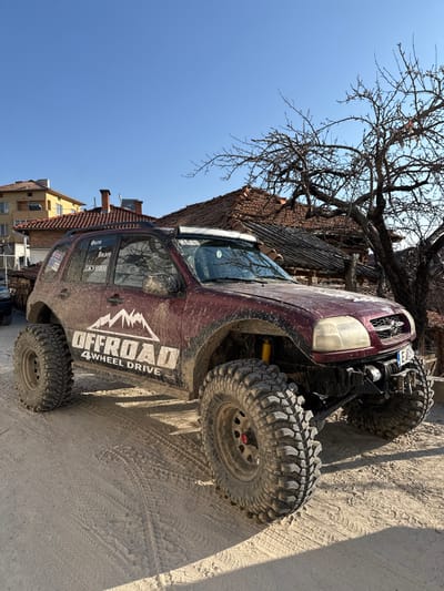 A heavily modified, dark red off-road SUV is parked on a dirt road in Breznitsa, Bulgaria, under a clear blue sky. The vehicle, appearing to be a Suzuki Grand Vitara or similar model, is covered in dried mud and features large, knobby off-road tires, a lifted suspension with visible yellow shock absorbers, a front-mounted winch, and a roof-mounted LED light bar. White decals on the side read "OFFROAD 4 WHEEL DRIVE" with a mountain graphic, and additional Cyrillic text on the rear passenger window states "Фото и Видео" (Photo and Video) and "ДЖУНИН" (DZHUNIN). A Bulgarian license plate is partially visible on the front bumper, beginning with "E 4...". The ground consists of compacted dirt with visible tire tracks. In the background, traditional houses with red tile roofs and chimneys are present, along with a multi-story building with a yellow facade. A prominent deciduous tree with bare branches stands to the right, casting shadows on the ground. No people are visible.