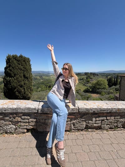 A full-length, rotated photograph captures a young woman smiling and posing on a stone wall, overlooking the scenic Tuscan countryside of San Gimignano, Italy. The woman has long blonde hair, wears sunglasses, a light-colored plaid shirt over a dark top, blue jeans, and brown sneakers with white laces. She is positioned horizontally in the frame, with her left arm outstretched as if reaching towards the distant landscape, and her body leaning back against the stone wall. Her bright, open smile conveys a sense of joy and relaxation, suggesting she is enjoying the moment and the view.

Behind her, the stunning backdrop features rolling green hills, lush vineyards, and numerous trees, including a prominent dark green conifer in the mid-ground. Part of a historic stone building or structure is visible in the upper background, characteristic of the region. The sky is a vast, clear blue, indicating bright, sunny weather. Strong shadows on the paved path and wall suggest the image was taken during midday or early afternoon. The immediate foreground includes the textured, ancient-looking stone wall she is resting on and a section of paved path. The unique rotation of the image gives a playful, almost gravity-defying feel to her pose, capturing a memorable moment amidst the beautiful Italian landscape. There is no visible text in the image.