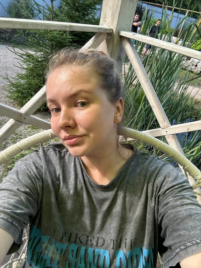 In Lyamino, Russia, a young woman with blonde hair pulled back is photographed from the chest up, seated in a hanging chair. She is wearing a dark grey t-shirt with a faded blue graphic. The background includes a wooden structure, tall green reeds, and two men in dark t-shirts standing among the reeds. The woman's face is turned towards the camera, with her head tilted slightly. Sunlight illuminates her face, creating a sheen on her skin. The environment suggests an outdoor setting, possibly a park or recreational area.