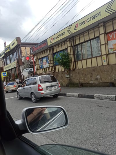 The image captures a street scene in Dariali, Georgia, featuring a silver station wagon stopped at a traffic light. The traffic light is red. A pedestrian crossing sign is visible on the left, indicating a crosswalk. The building in the background has a sign that reads "Ре: Старт" (Re: Start). There are other vehicles and people in the vicinity, suggesting a moderately busy urban environment. The sky is overcast. The photograph is taken from inside another vehicle, as evidenced by the reflection of a hand in the side mirror.