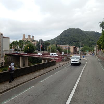 The image captures a view of the city of Foix, France, with the prominent Château de Foix visible on a hill in the background. In the foreground, a person wearing a grey hooded sweatshirt and a backpack is walking along a sidewalk next to a road. A red-railed bridge spans a river or ravine, with several vehicles, including white vans and a blue car, crossing it. The road itself has white and green lane markings. The overall scene is characterized by overcast skies and a mix of urban architecture and natural landscape, including a densely forested mountain in the distance. Buildings of various architectural styles are present, including apartment complexes and older structures. Yellow and red flags are visible on poles along the roadside. The lighting suggests it is daytime.
