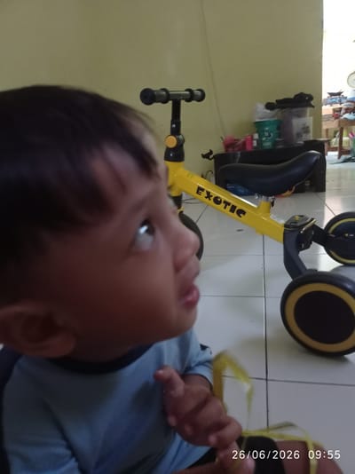 A close-up, eye-level shot shows a young child looking upwards and to the right, their face illuminated by soft indoor lighting. The child has dark hair and is wearing a blue t-shirt. Their gaze is directed towards something out of frame, with a hint of curiosity or wonder. In the background, a bright yellow children's balance bike with black wheels and seat is visible. The word "EXOTIC" is printed in black on the bike's frame. The bike appears to be in a room with tiled flooring and a light yellow wall. Behind the bike, a shelf holds various items, including containers and bottles, suggesting a home environment. Further in the background, a doorway leads to another room where furniture and items are dimly visible. A timestamp in the lower right corner reads "26/06/2026 09:55". The overall scene suggests a casual moment at home in Sukaraja, Indonesia, likely in the morning.