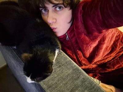 A person in a reddish-purple fuzzy jacket pouts at the camera, their face close to a black and white cat lying on their lap. The cat's head rests on a grayish-green surface, possibly a leg covered in denim. The person has dark hair and light eyes. The photo seems to be a close-up selfie, giving a personal and intimate feel.