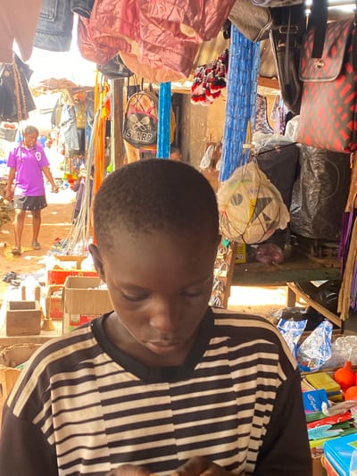 The image depicts a street market scene in Amaseri, Nigeria. In the foreground, a young boy with dark skin and short hair is looking downwards, focused on something in his hands. He is wearing a black and white striped t-shirt. The background reveals a busy market with various stalls displaying goods. To the left, a person in a vibrant purple t-shirt and black shorts is visible, seemingly interacting with the market environment. Various items are hanging from the stalls, including bags, textiles, and what appear to be plastic items. Several cardboard boxes and plastic containers are also present, holding merchandise. The ground is dirt and littered with some debris. The overall impression is of a lively and functional marketplace.