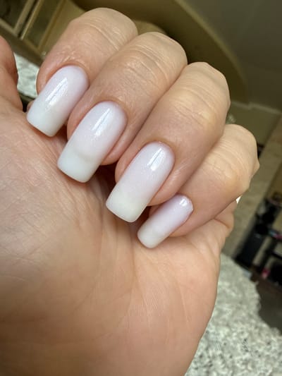 A close-up shot focuses on a person's hand with neatly manicured square-shaped fingernails. The nails are coated in a milky white polish with a subtle shimmer. The skin of the hand appears smooth with visible creases on the knuckles and fingers. The background is softly blurred, suggesting an indoor setting with warm lighting and some indistinct objects, possibly furniture or shelves, visible on the right side. The image does not contain information about the city of Magadan, Russia.